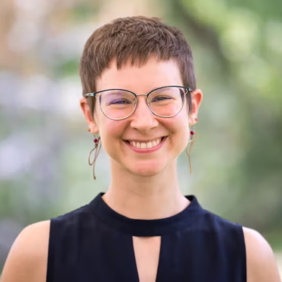 Gillian Esquivia-Cohen, a white woman with close-cropped brown hair, glasses, and dangly earrings, smiles at the camera. She wears a black shirt with keyhole cutout and stands in front of a background of foliage.