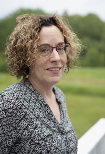 Debra Spark, a white woman with brown curly hair and glasses. She is wearing a black-and-white patterned shirt and is standing outside with a white railing, a field, and trees blurred behind her.