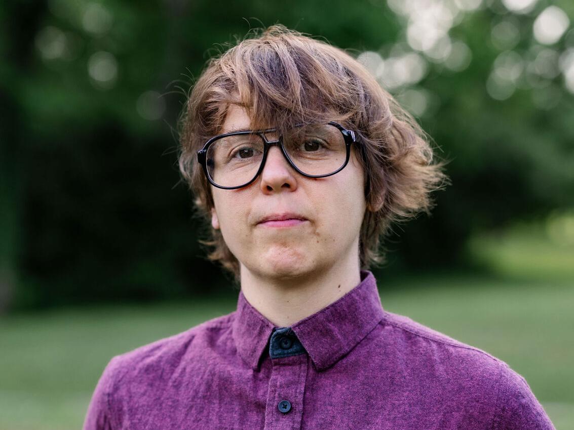 Lydi Conklin, a white person with a Beatles-style haircut wearing aviator-type glasses and a purple button-down shirt, looks into the camera in front of some greenery. They half-smile.