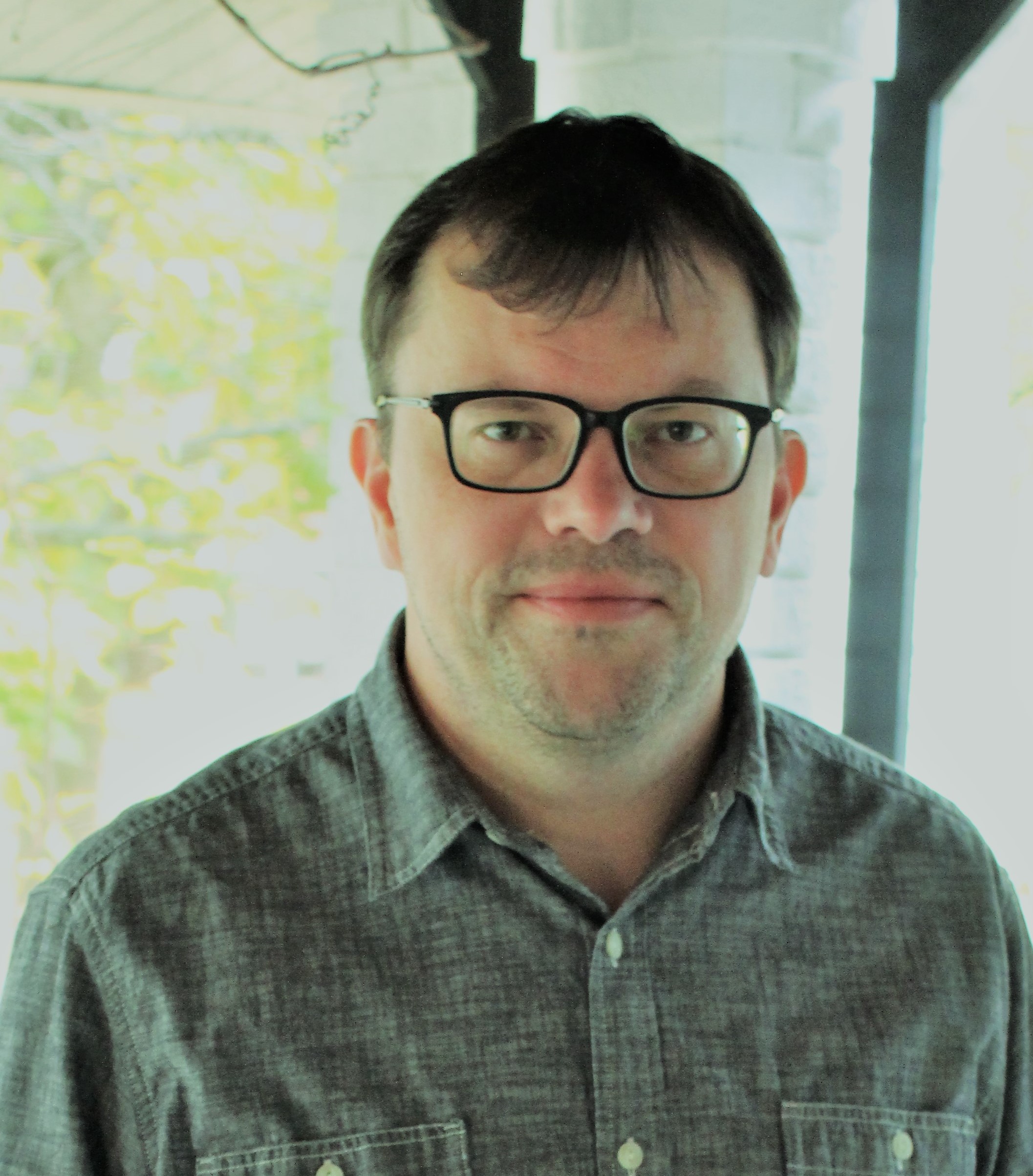 Chris Haven, a white man with black-framed glasses, wears a button-up shirt and stands in front of a window.