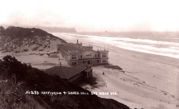 Period photograph of Bayocean Natatorium from a cliff overlooking the beach. It's a turn-of-the-20th-century white building with curved roof and covered porch. To its right is the ocean, and to its left are a series of dunes covered with brush.