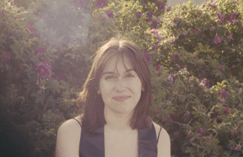 Bella Gibb, a white woman with brown shoulder-length hair and a sleeveless shirt. She is sitting in front of a bush with purple flowers, and the sun is causing a slight lens flare above and to the left of her head.