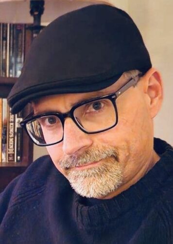 Mark Strohschein, a white man with glasses, a gray goatee, and a black flat cap. he's wearing a dark sweater and in front of a bookcase and cream-colored wall.