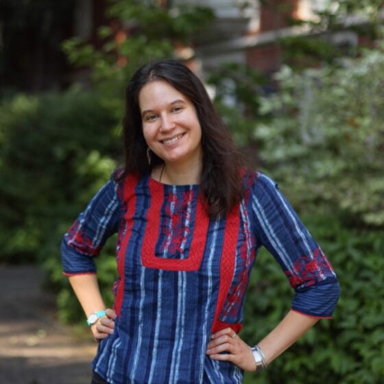 Zoe Ballering, a hispanic/white woman, stands in front of some foliage wearing a blue and red peasant shirt. She has her head cocked to the left and smiles at the camera.