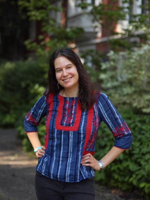 Zoe Ballering, a latina/white woman with dark brown shoulder-length hair, stands in front of some greenery with her head cocked to the left. She wears a blue-and-red peasant shirt and smiles warmly.