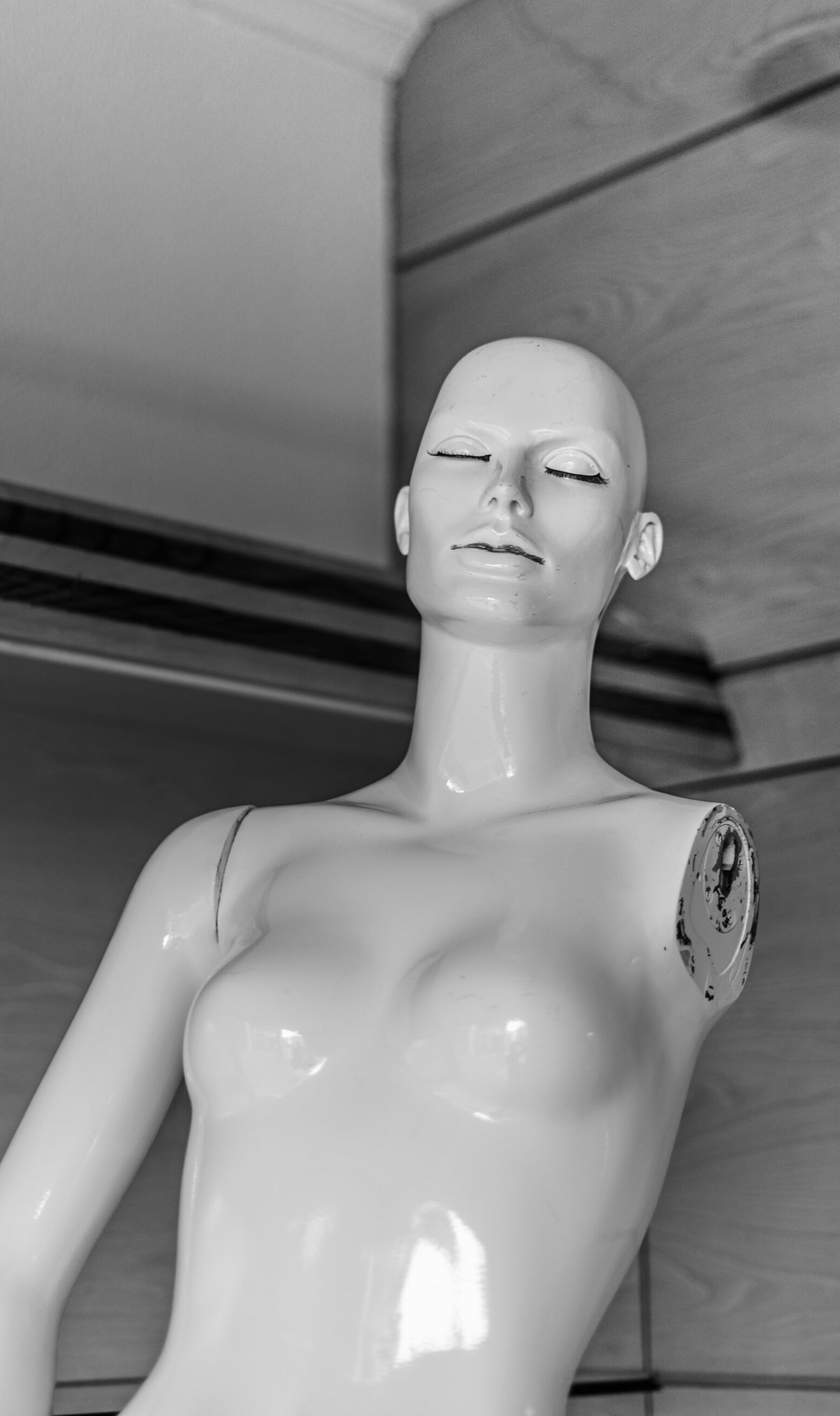 A bald, shiny white mannequin of a woman's torso. It is missing a left arm and its eyes appear to be closed. It rests on a gray background.
