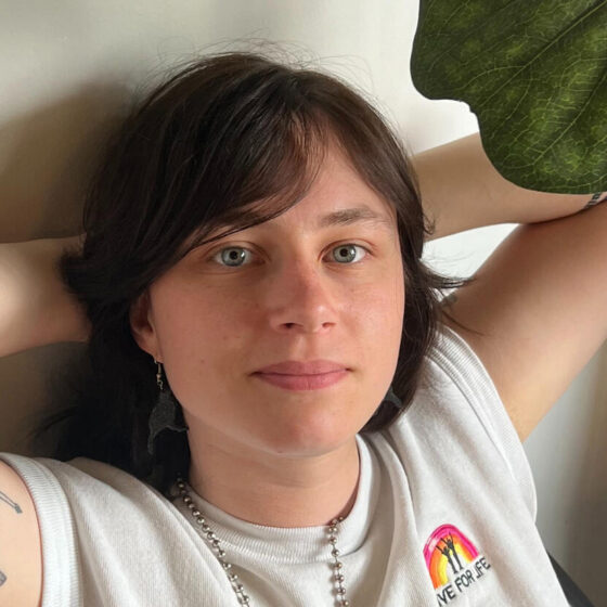 Gabrielle Grace Hogan, a white person with brown hair wearing a white tee shirt, has their arms crossed behind their head and leans against a white wall. A green leaf from a houseplant is showing, top right.