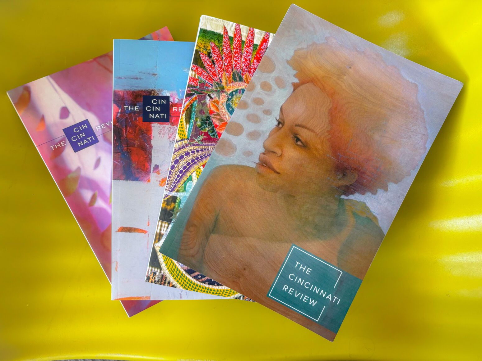 A selection of print copies of CR fanned out on a wavy lime green background. The top cover features a watercolor painting of a Black woman with orange hair. The painting has a slightly aquatic quality to it.