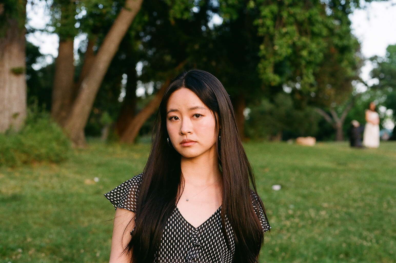 Ting Lin, an Asian woman with long brown hair wearing a black v-neck shirt, stands in a park in front of some palms and cocks her head to the left. She does not smile.