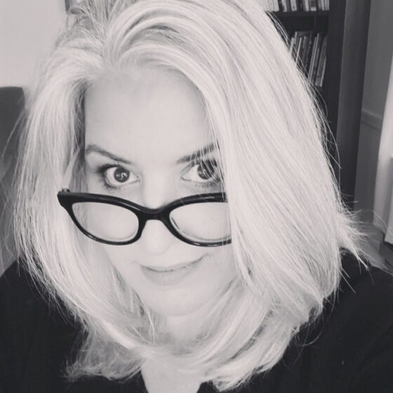 Suzanne Frischkorn, a latina woman with white hair, peeks over black-rimmed glasses and smiles. The photo is black-and-white.