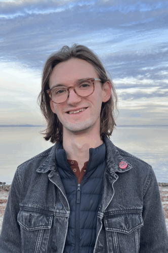 A white man with brown hair to his shoulders and tortoiseshell-frame glasses. He's wearing a black jean jacket over a puffer jacket and shirt, and he's standing on a beach in front of a body of water.