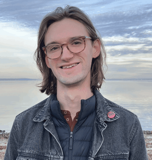 A white man with long light-brown hair. He's wearing glasses and a jean jacket and standing in front of a body of water with clouds in the sky