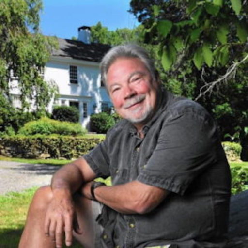 Robert Cording, a white man with short gray hair and gray beard, sits casually with an arm on his knee in front of an idyllic white house surrounded by greenery.