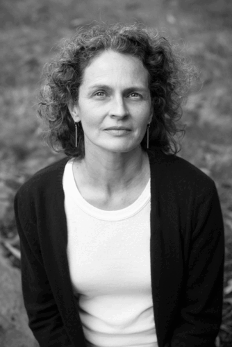 A black-and-white photo of a white woman with curly hair. she is wearing a dark sweater and light-colored shirt and sitting oustide.
