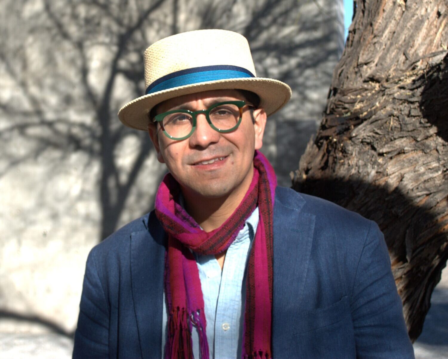 Edgar Garcia standing in front of a tree. Garcia is wearing a brimmed hat, glasses with green frames, and a pink scarf.