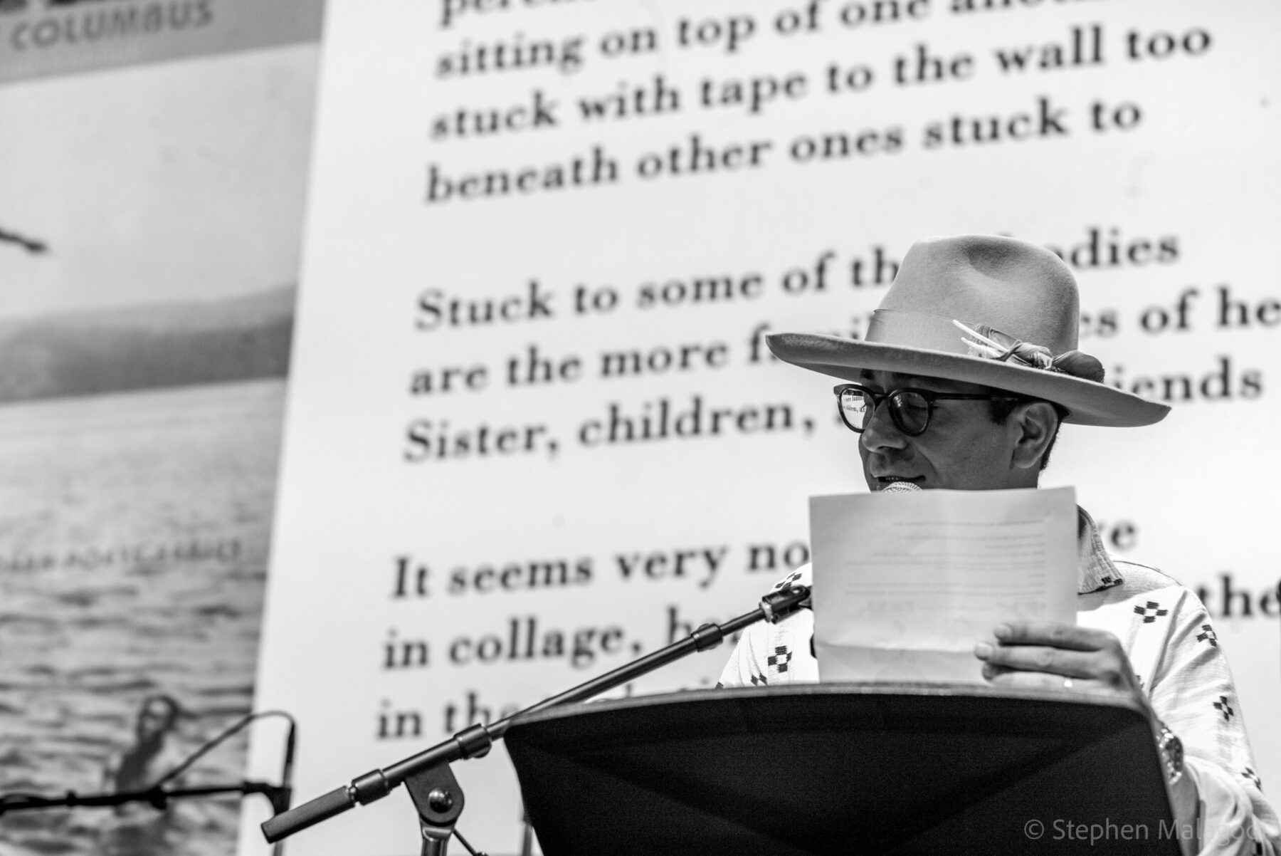Edgar Garcia speaks into a microphone behind a podium and in front of a banner with one of Garcia's poems. Garcia is wearing a brimmed hat and glasses.