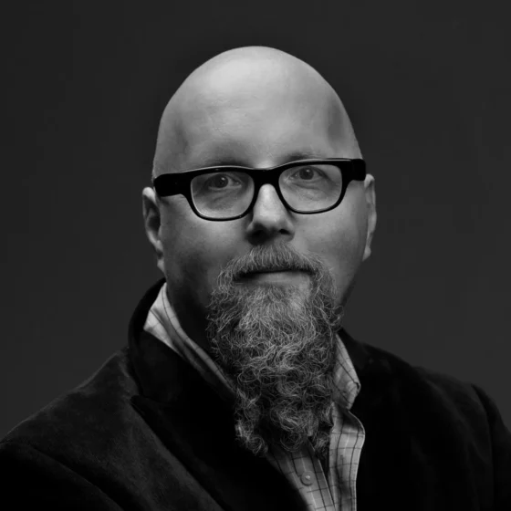L.S. Klatt, a bald white man with a long, curly goatee, wears dark-rimmed glasses and a half-smile. The photo is black-and-white.