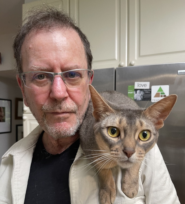 David Lazar, a white man with gray hair and short beard and mustache, poses with a curious cat on his shoulder. The cat looks like some kind of fancy purebred with short hair and bright yellow eyes. It stares directly into our souls.