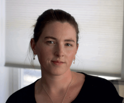A Palestinian American woman with brown hair and hazel eyes. She's wearing a dark shirt, dangling earrings, and a necklace chain, and standing in front of a white window shade.