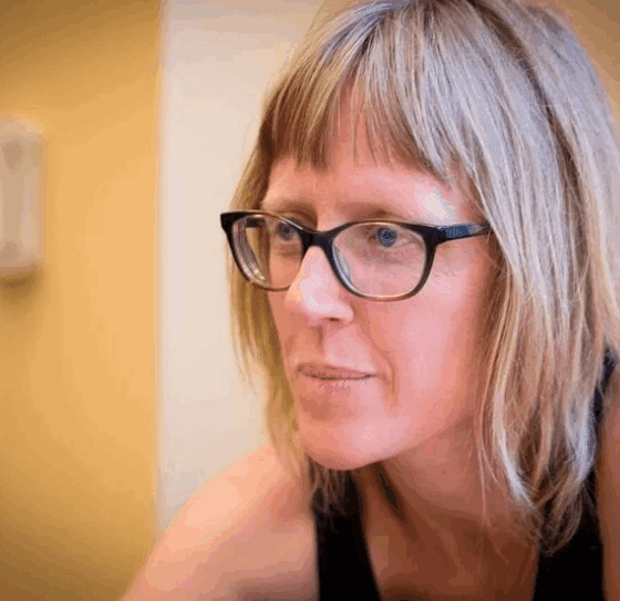 Sarah Viren, a white woman with a blond bob with bangs and dark-framed glasses, looks into the middle distance in front of a yellow wall.