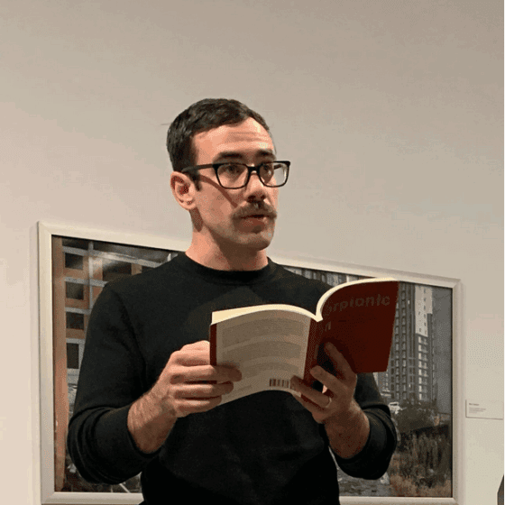 Conor Bracken, a white man with dark mustache and short dark hair, reads aloud from a book. He wears dark-rimmed glasses. He stands in front of an artwork in a gallery space.