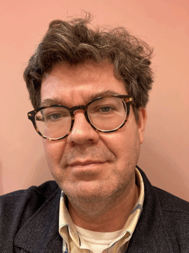 Matthew Thorburn, a white man with brown and gray hair, and tortoiseshell glasses. He is wearing a gray tweed jacket and colored shirt and standing in front of a warm pink wall.