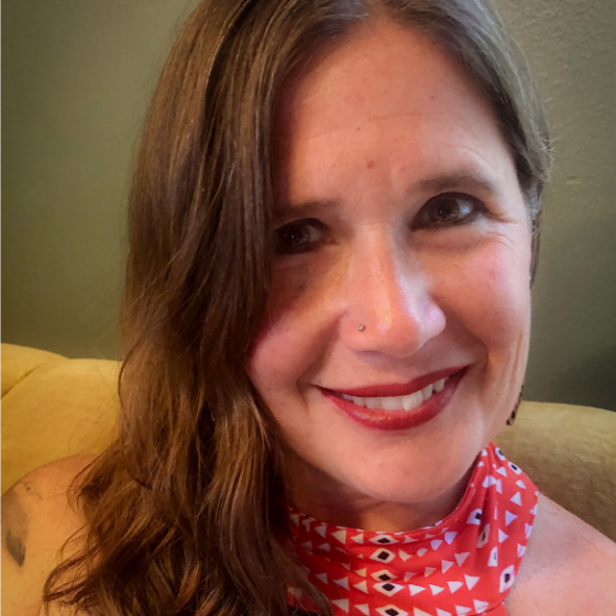 Alexandra Teague smiles while sitting on yellow furniture in front of a sage colored wall. She wears red lipstick, eyeliner, and has a stud nose piercing.