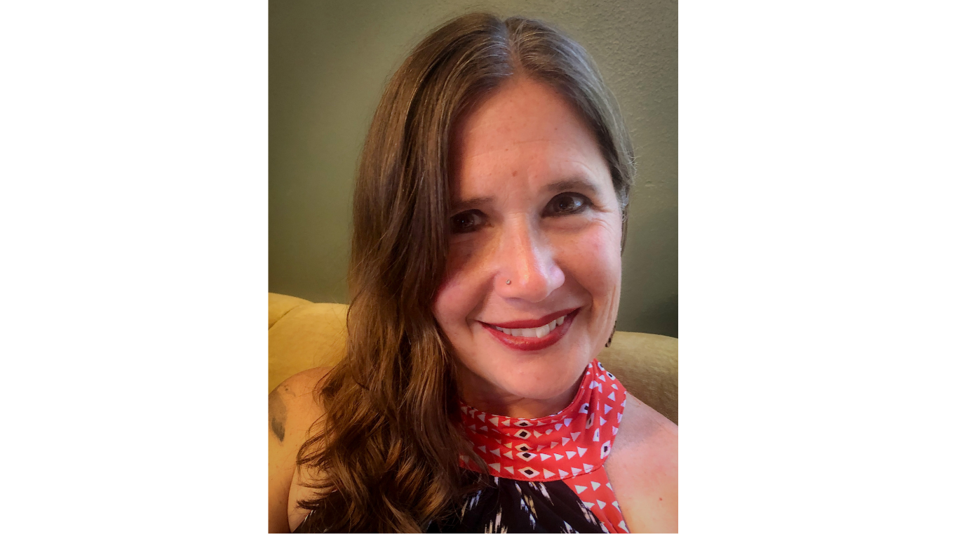 Alexandra Teague smiles while sitting on yellow furniture in front of a sage colored wall. She wears red lipstick, eyeliner, and has a stud nose piercing.
