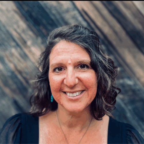 Maya Jewell Zeller, a white woman with brown eyes and gray/dark hair. She is smiling, wearing blue earrings and a necklace, and standing in front of a textured wood