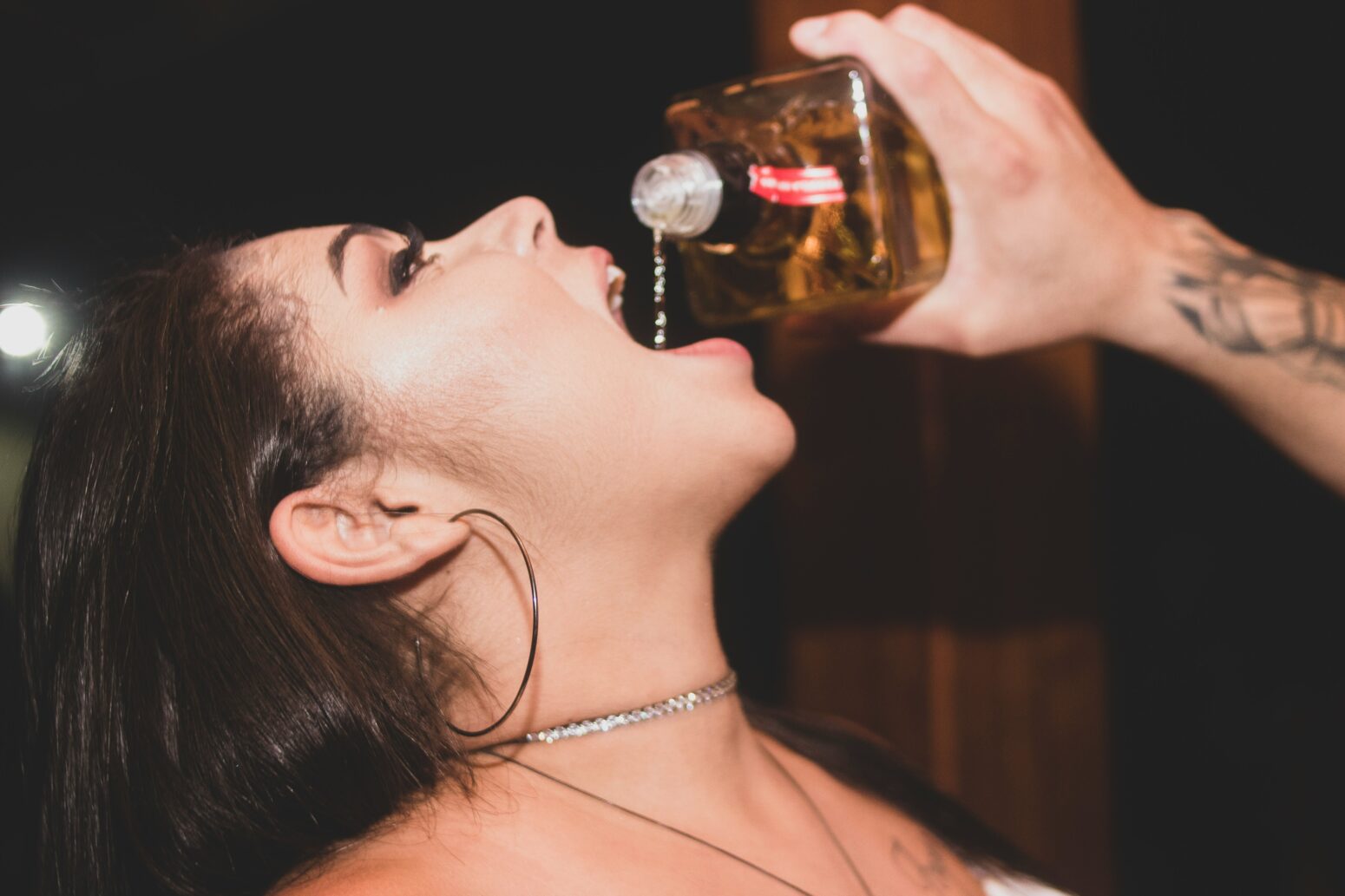 A white male hand pours alcohol straight from the bottle into the mouth of a woman with large hoop earrings and pink eyeshadow.