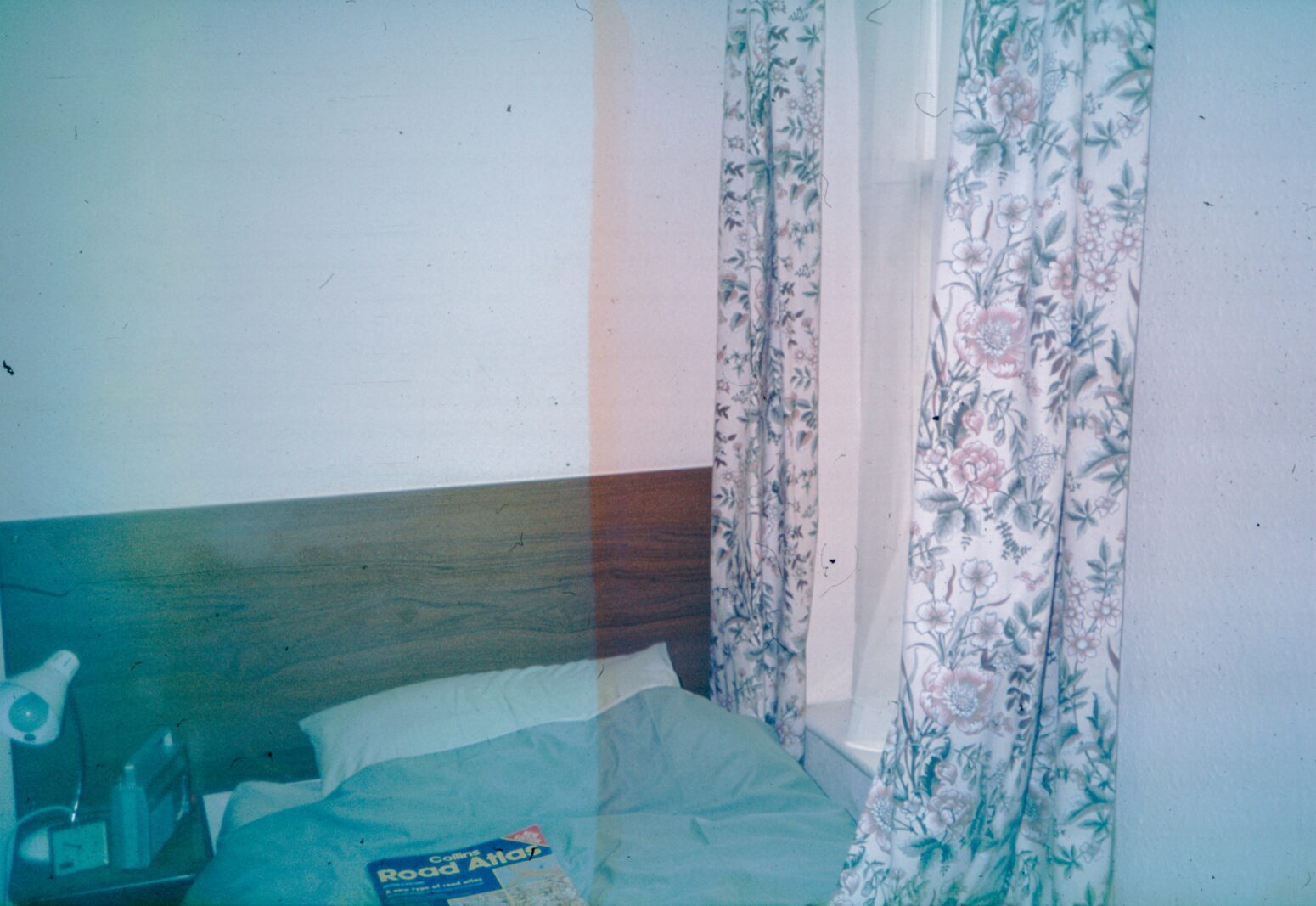 A film photo, with distortion, of a 1980s bedroom with faux wood headboard and floral curtains. A road atlas sits on the bed. There is a lamp and radio on the side table.