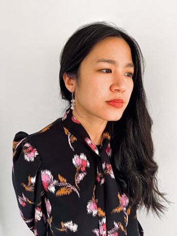 Marianne Chan, an Asian-American woman, stands in front of a white background. She has long dark hair draped over her left shoulder and wears dangly earrings and a black secretary blouse with large pink flowers on it. She glances away from the viewer.