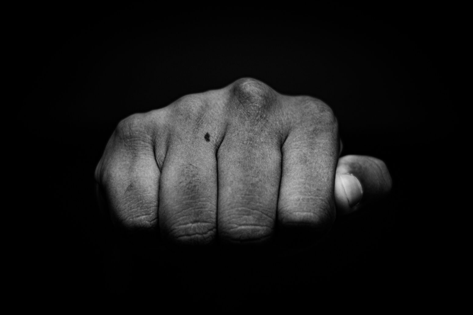 Finger-and-knuckle-view of a dark-skinned fist, from the perspective of one who's about to get punched. There is a birthmark on the third knuckle. The photo is taken in black-and-white.