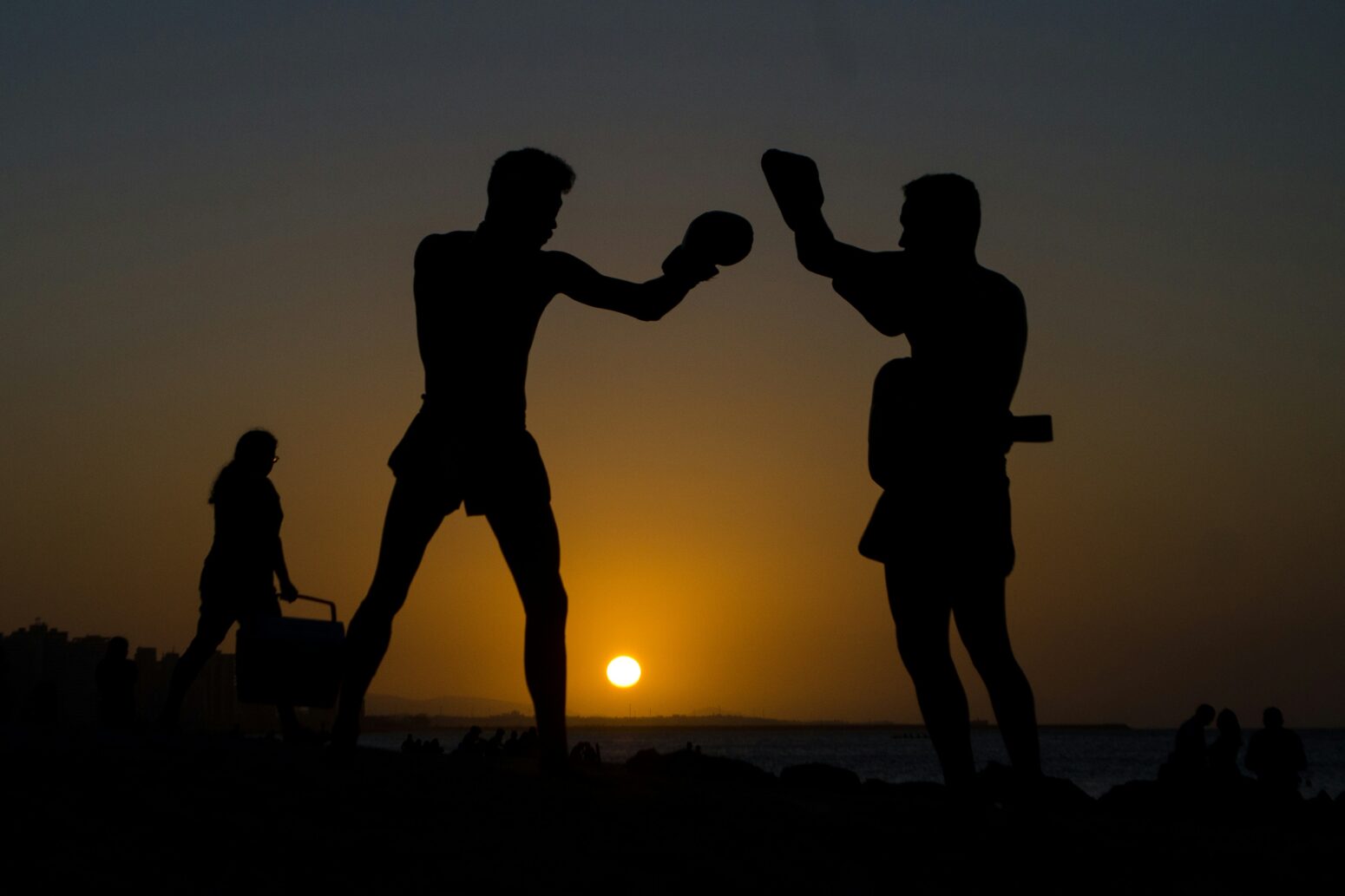 Two male silhouettes box each other against a sunset. Another silhouette in the distance pushes some kind of cart.
