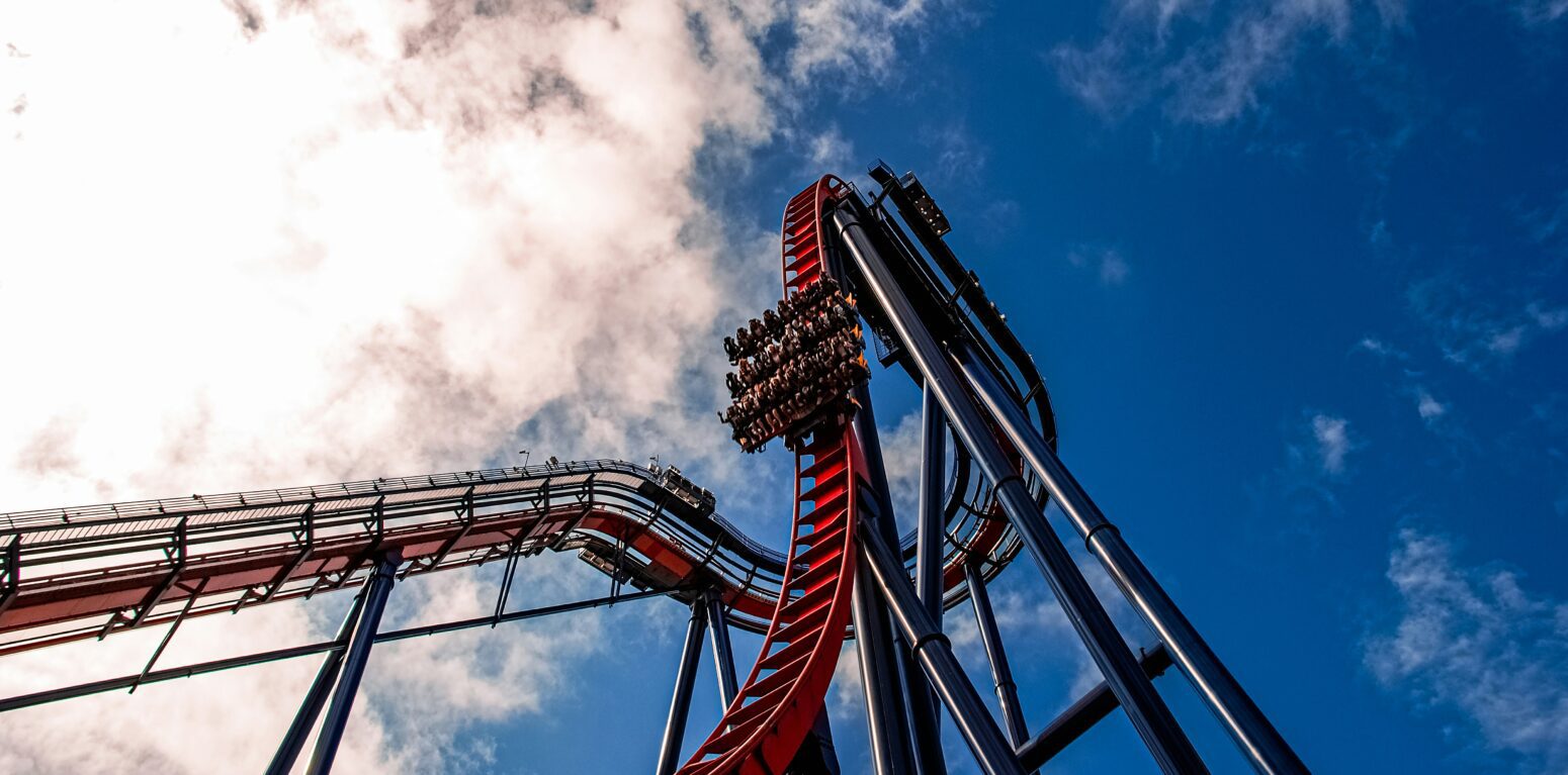 The sheiKra roller coaster at Busch Gardens, showing riders descending from the top of a drop. Blue sky with fluffy clouds is above them. The tracks of the coaster are red.
