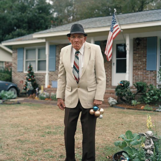 An old white man with a grizzled face stands in his close-cropped front yard in front of a midcentury bungalow. He wears a tan blazer, red, gray, and blue striped tie, brown fedora, and brown pleated pants. He is slightly hunched. In his left hand he holds a clutch of Christmas ornaments. Some of the bushes near his house are lit with multicolored fairy lights. An American flag hangs in front of his house.