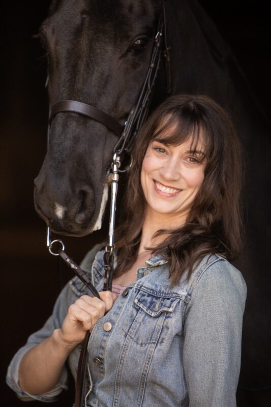 Emma Hudelson, a white woman with shoulder-length brown hair with bangs, stands in front of a black background holding the reins of a black horse with a white star on its nose. It peers over her shoulder right at us. Hudelson wears a denim jacket. She smiles at us.