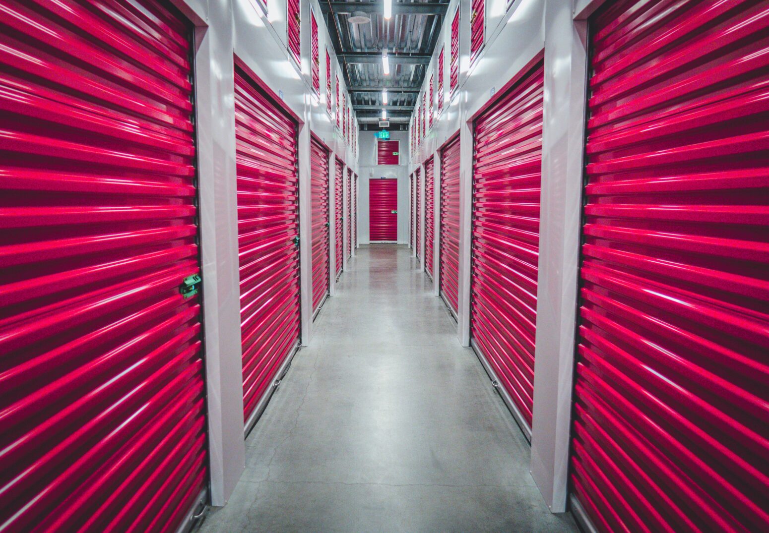 The hallway of an indoor self-storage center. The corrugated metal doors are red while the walls and concrete floor are gray. The hallway is lit by harsh fluorescents.