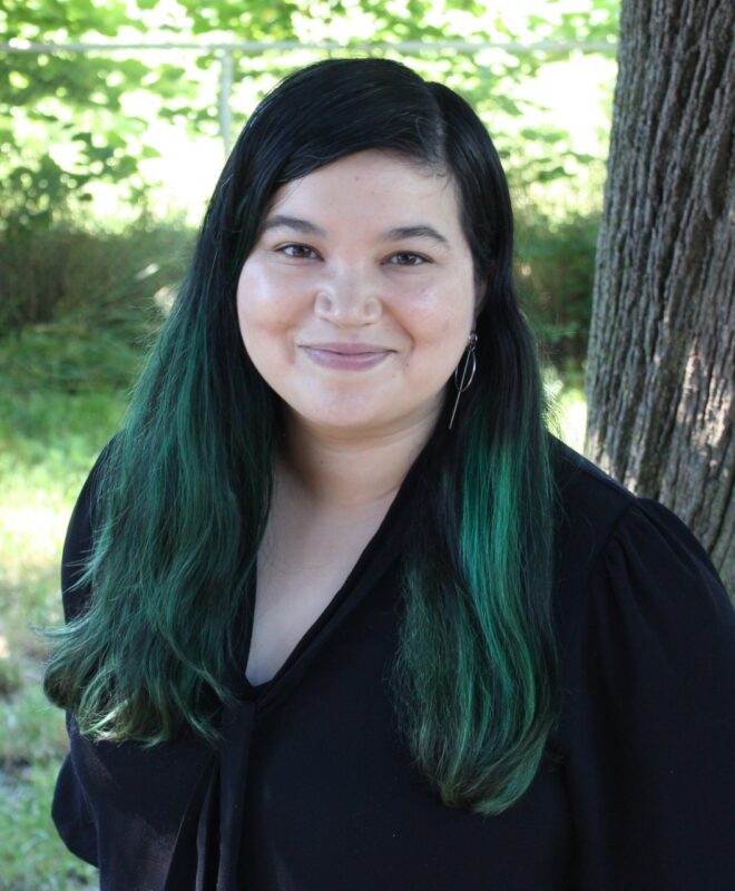 Maggie Su, an Asian-American woman, smiles up at the camera. Her long, dark hair has streaks of green and she wears a black blouse. She stands next to a tree in a wooded setting.
