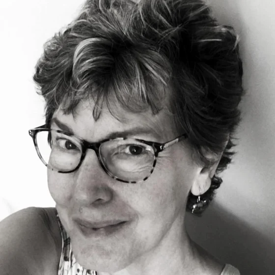 Mary Cisper, a white woman with a pixie cut wearing tortoiseshell glasses, smiles up at the camera in 3/4 profile. The photo is black-and-white.