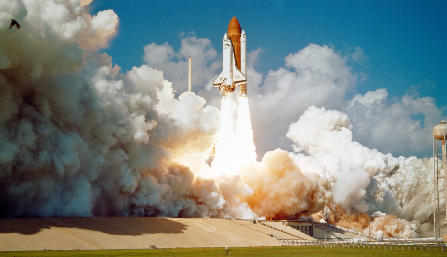 A space shuttle launches at Cape Canaveral. Fiery columns shoot out of the bottom of the rocket, with clouds of smoke enveloping the area. The sky above the rocket is blue.