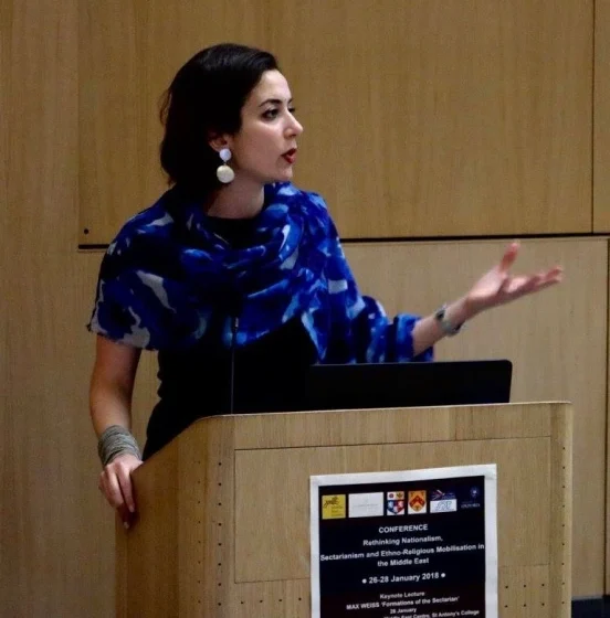Loubna El Amine, a Middle-Eastern woman wearing a blue dress, gold dangly earrings, and a cuff bracelet, speaks at a lectern in a wood-paneled room.