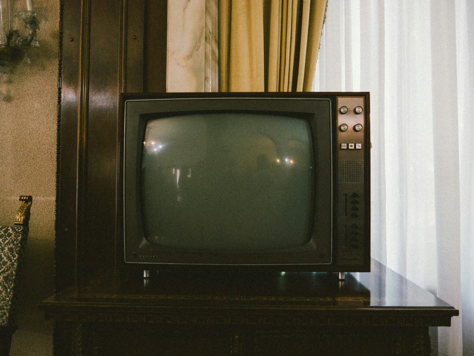 Late 80s/early 90s TV on a dark wood table in a dark wood paneled room. Gold curtains are pulled back behind the TV. The window behind it is covered with sheers.