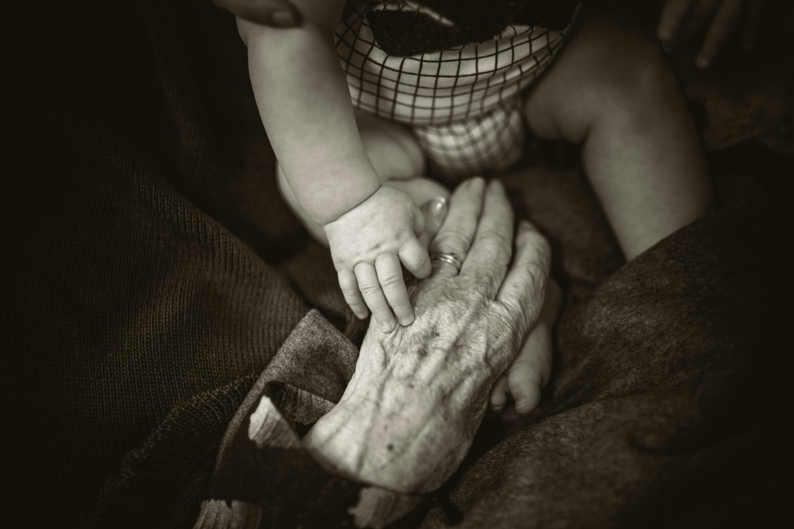 A chubby baby in a onesie, its face hidden, lays a hand on an old woman's hand. The elder's hand is lined and creased with age. It wears a wedding ring.