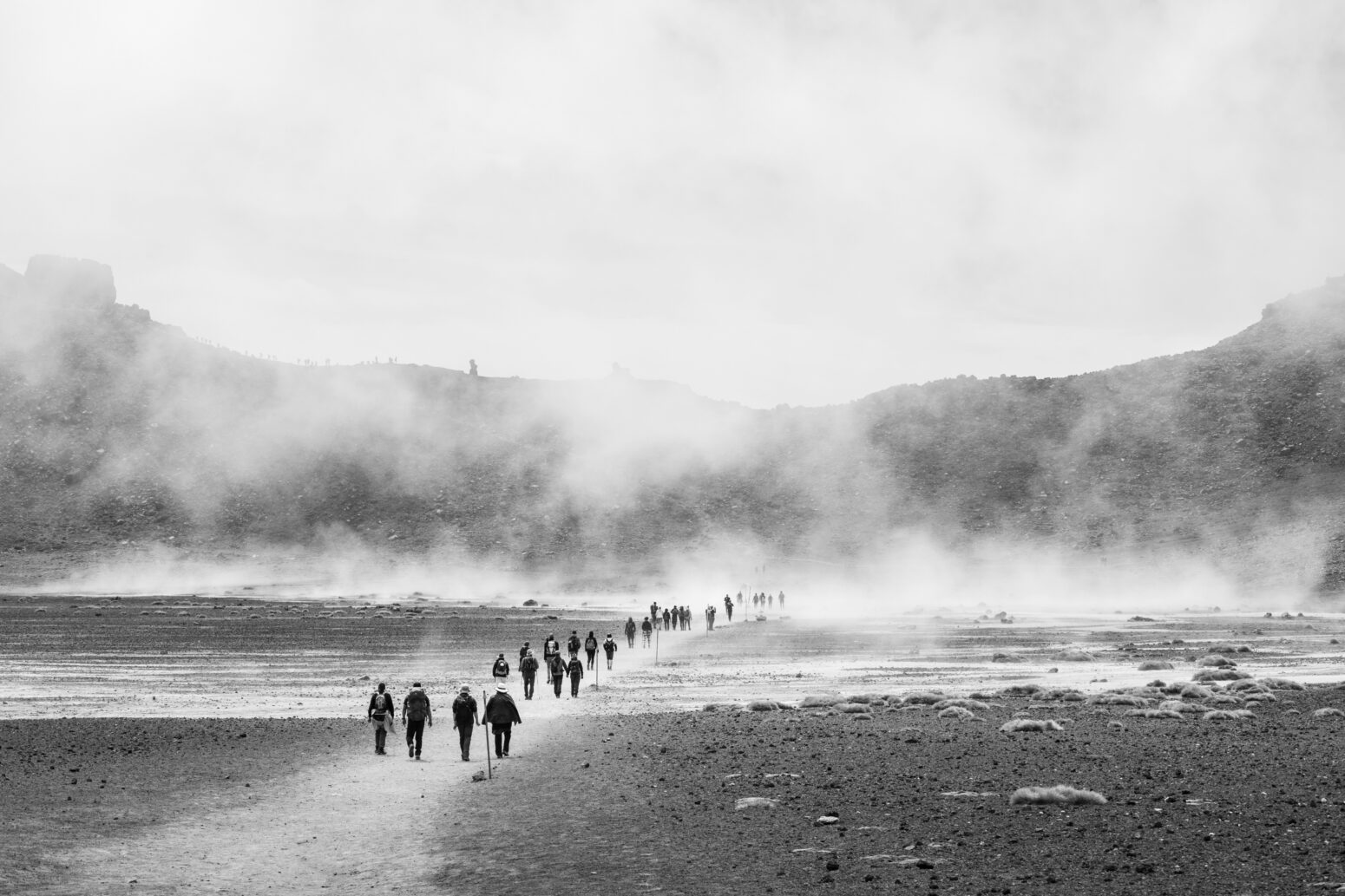 A long line of people walk toward a misty mountain. The photo is black-and-white.
