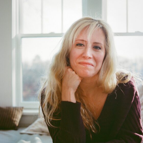 Leah Umansky, a white woman with long blond hair and bangs that fall across her forehead, wears a brown sweater and rests her chin on her fist. She smiles. She is sitting in a bright room, in front of a set of sash windows.