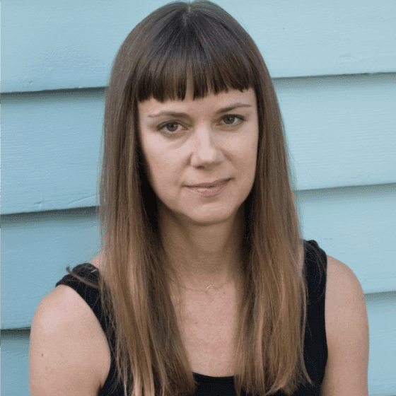 Leslie Brack, a white woman with straight brown hair and bangs, wears a black tank top and sits in front of a white clapboard wall.