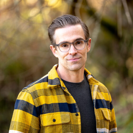 Brian Trapp, a white man with brown hair wearing glasses and a yellow-and-black buffalo check flannel shirt over a black tee, stands in front of some foliage and half-smiles.