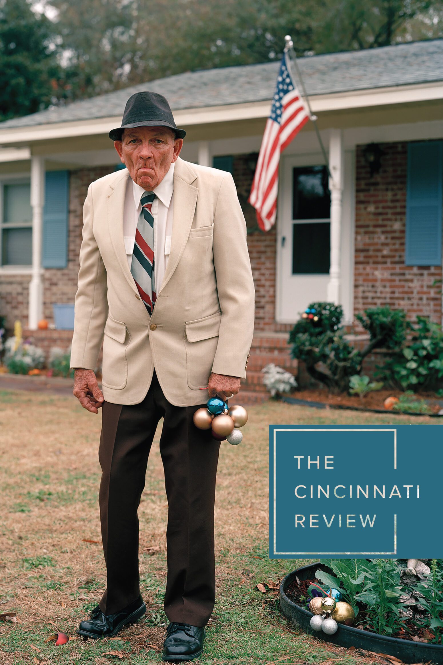 Cover of issue 22.2. An old man in tan jacket, brown pressed pants, gray fedora, and blue, red, and white tie holds a cluster of Christmas ornaments in blue and gold. He stands in his front yard, beside a patch of flowers. His house is a midcentury brick ranch with blue shutters. An American flag flutters in the breeze by the door.