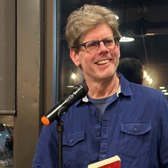 Matt Cashion, a white man with salt-and-pepper hair wearing glasses and a blue flap-pocket shirt, holds a book and stands in front of a mic. He smiles at someone in the crowd.